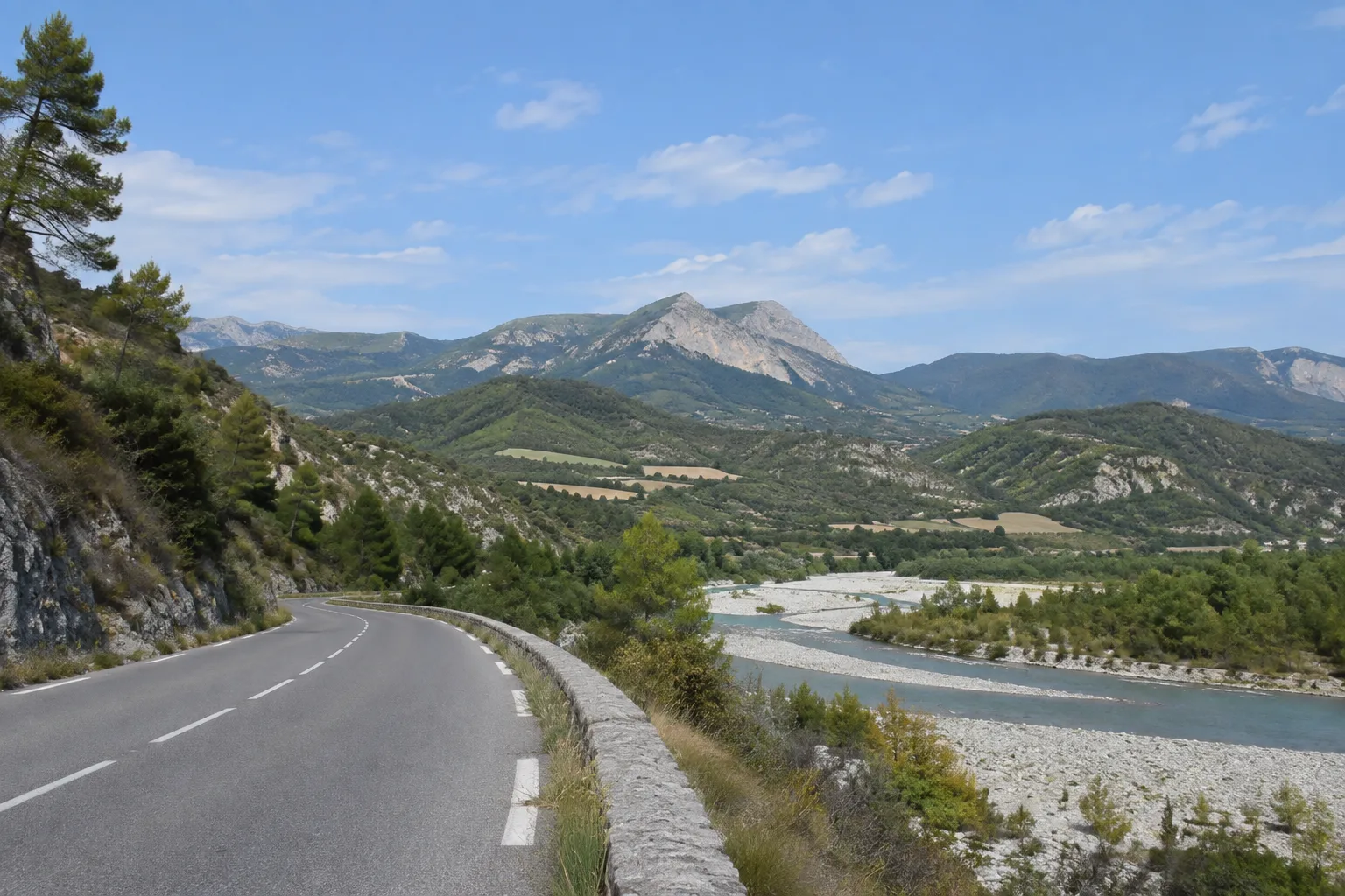 Une route suit le fond de la vallée de la Bléone entre la rivière et les versants.