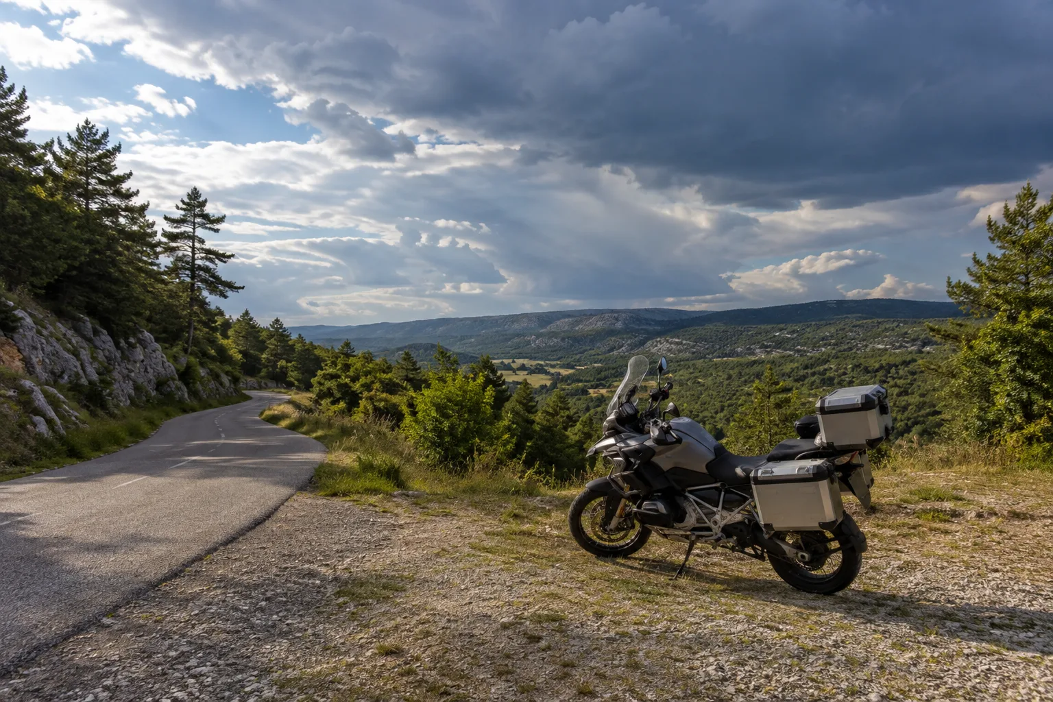 Une moto à l’arrêt sur un emplacement dégagé, près d’une route traversant un plateau sous un ciel nuageux.