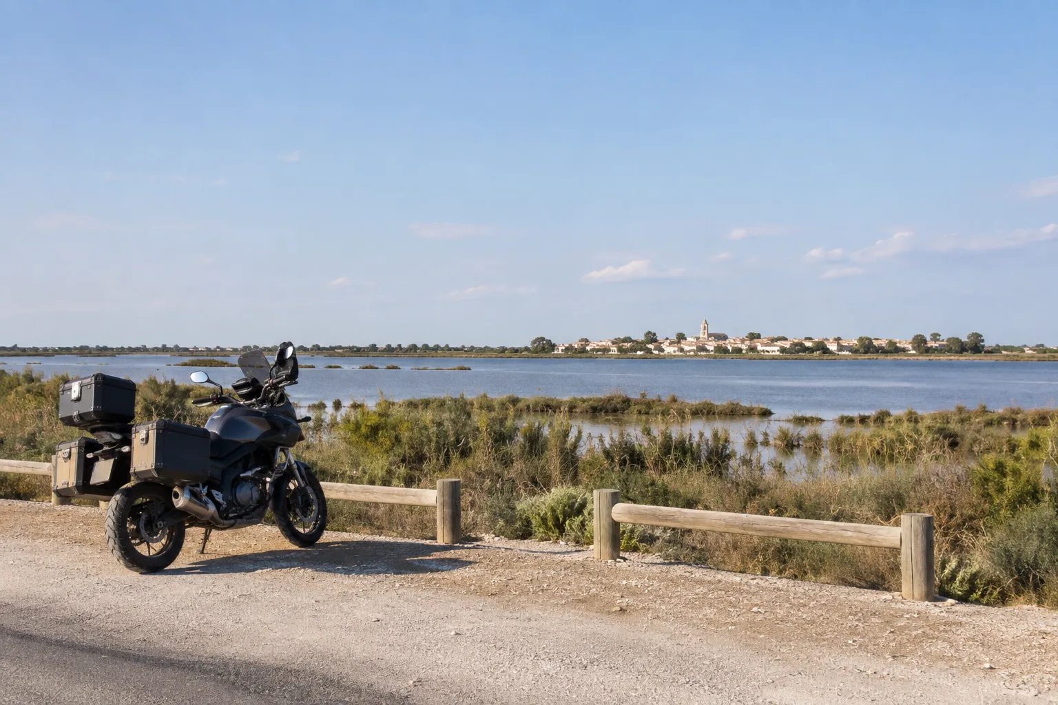 Une moto stationnée près d’une route bordant un étang, avec un village visible au loin.