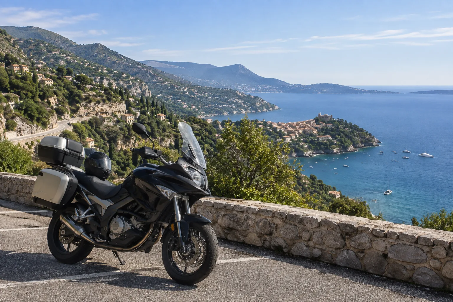 Une moto stationnée au-dessus de la Méditerranée, avec une route en corniche et un village perché en arrière-plan.