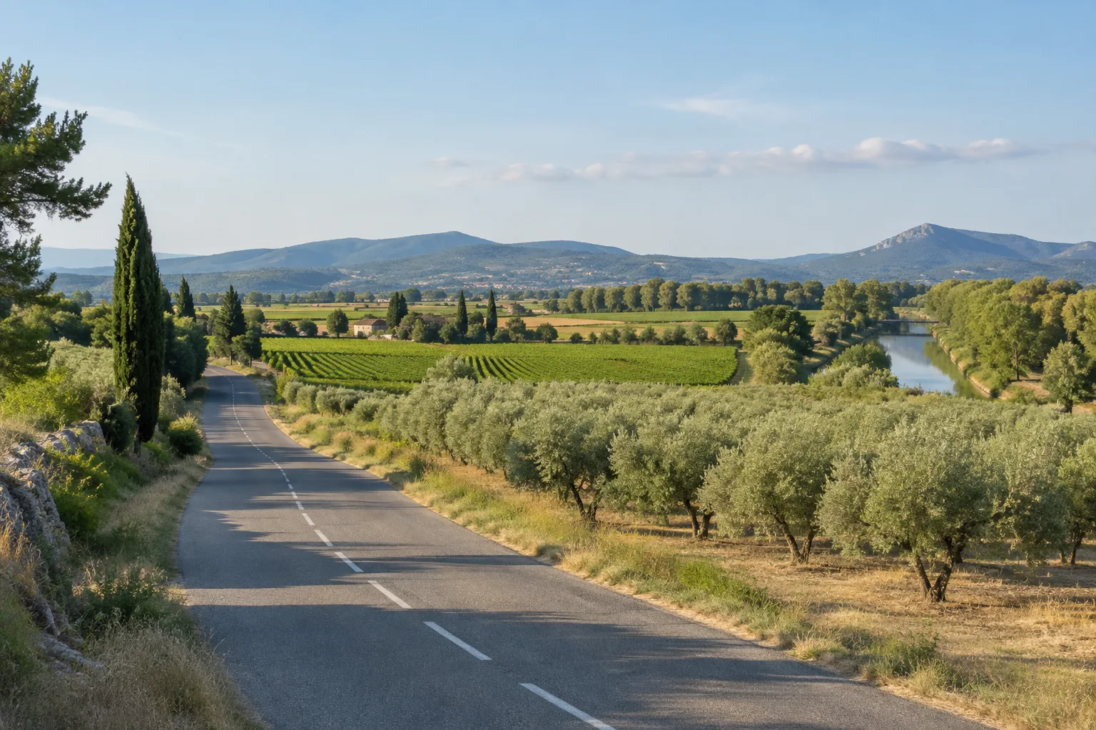 Une route traverse une plaine provençale cultivée, avec un canal bordé d’arbres et des collines en arrière-plan.