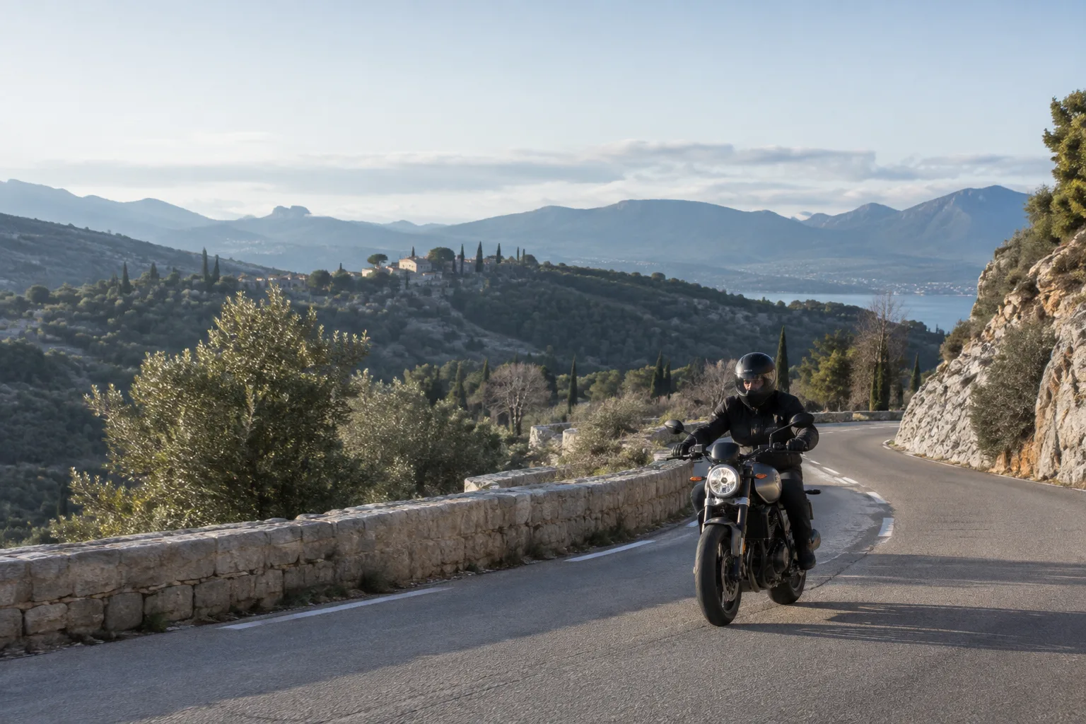 Un motard roule sur une route bordée de collines provençales sous une lumière hivernale.