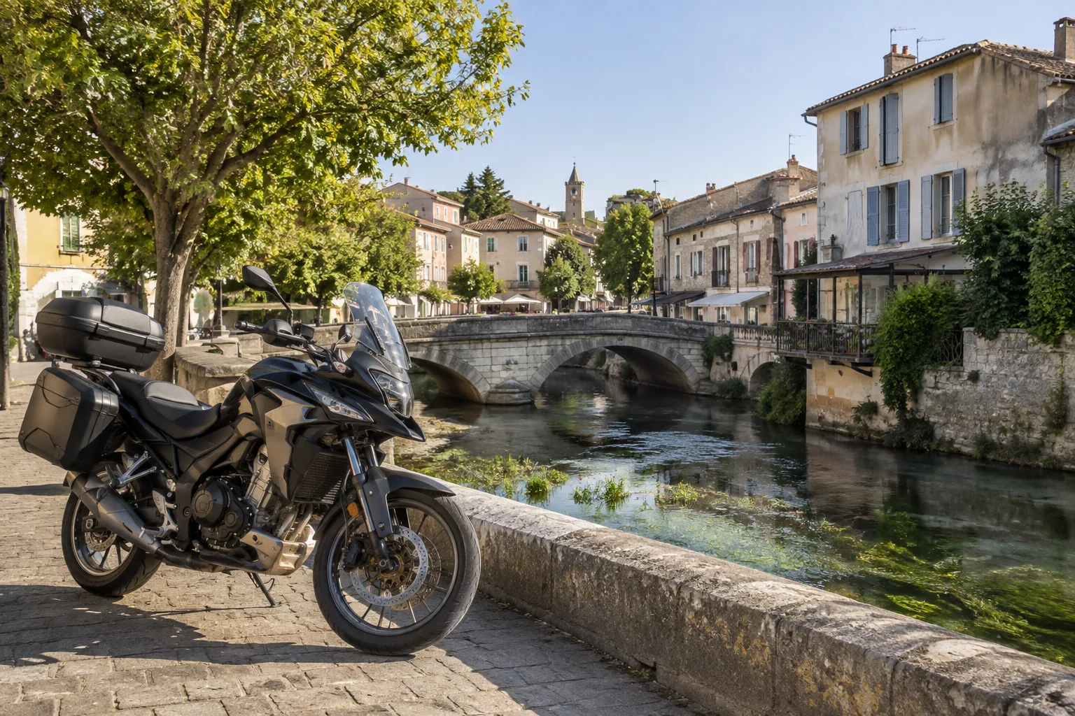 Une moto stationnée près d’un pont de pierre au-dessus de la Sorgue, devant des maisons provençales.