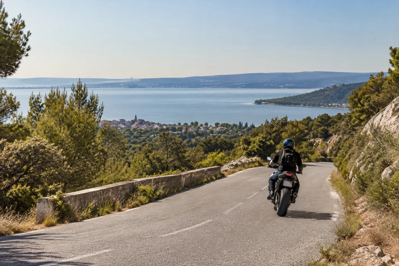 Un motard roule sur une route bordée de végétation provençale, avec l’étang de Berre visible au loin.