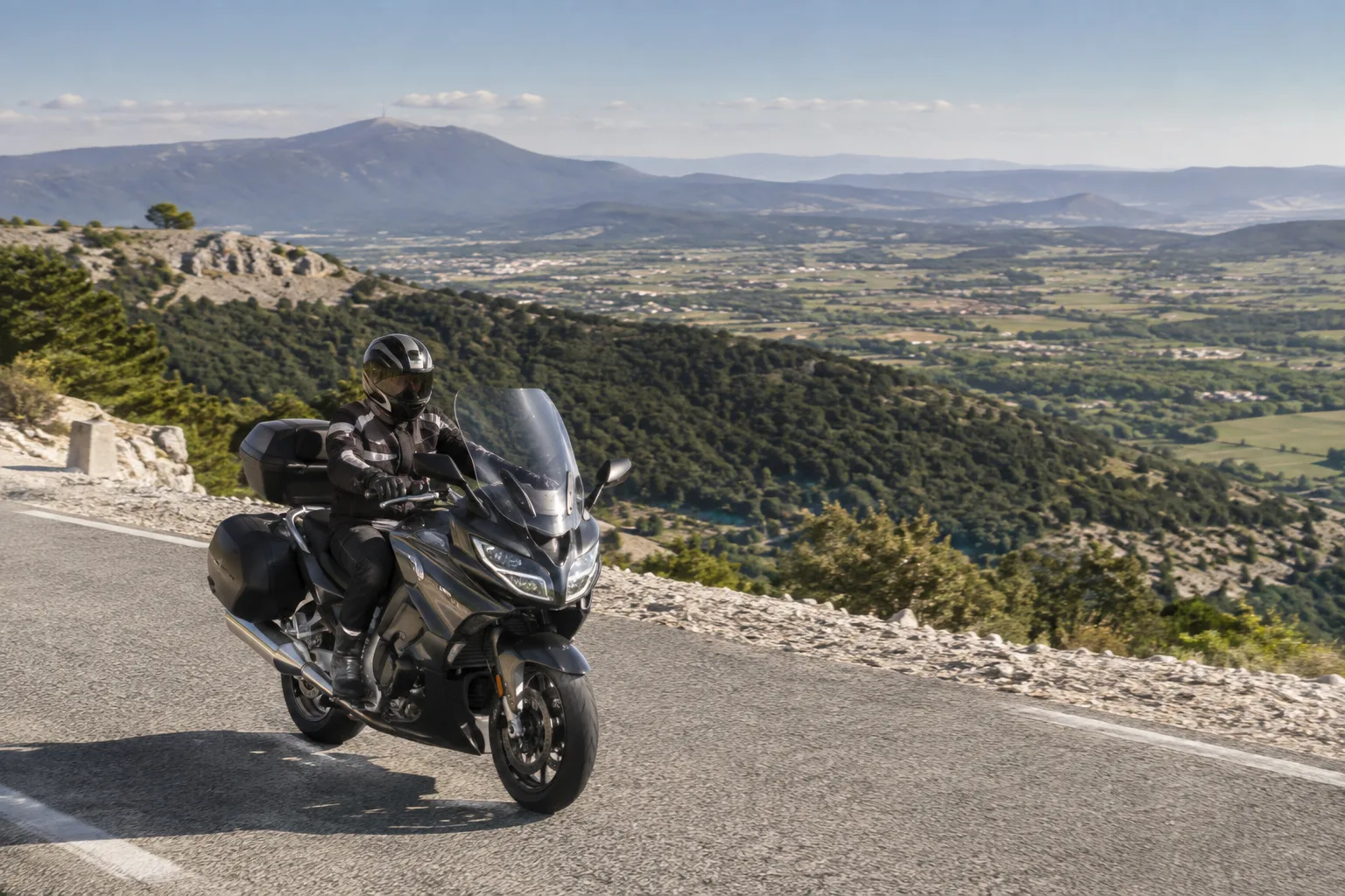Une moto roule sur une route sinueuse du Ventoux, entre forêt et reliefs provençaux.