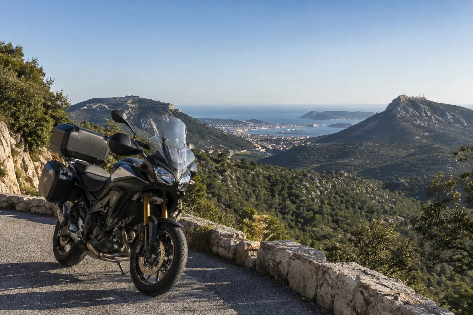 Une moto stationnée sur une route en hauteur, avec les collines du secteur de Toulon et la rade au loin.